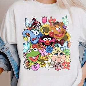 Może przedstawiać: Biała bluza z okrągłym dekoltem z kolorową grafiką postaci z Muppetów, w tym Kermita Żaba, Miss Piggy i Fozzie Bear, otoczonych kwiatami i motylami. Projekt jest w stylu kreskówkowym.