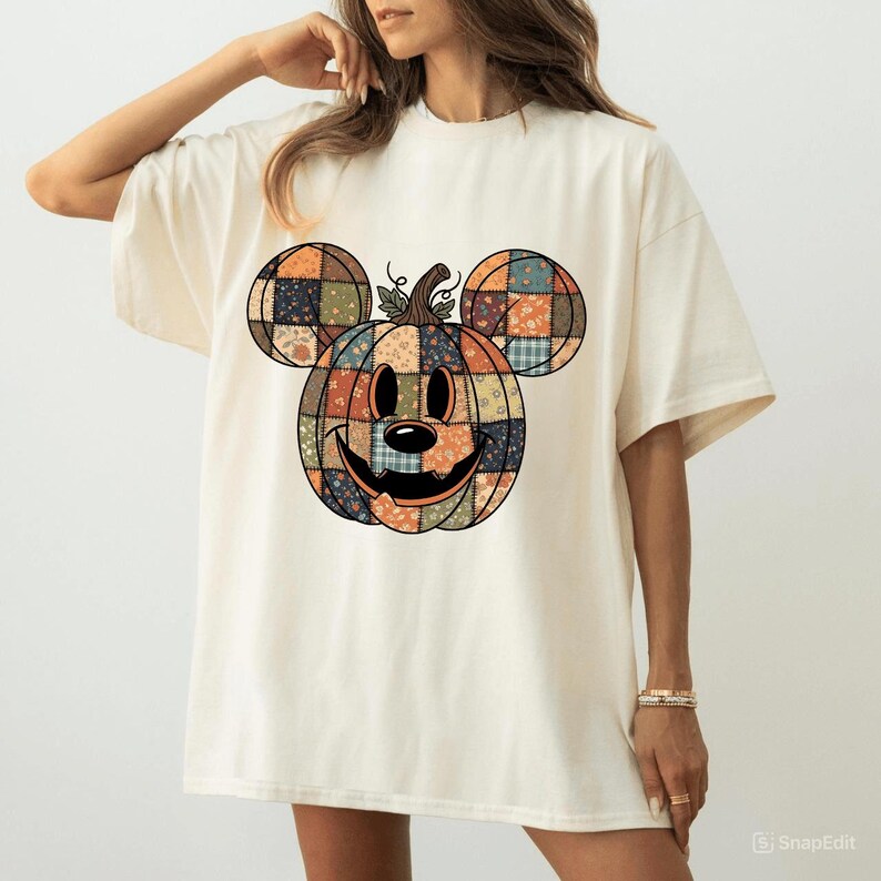 May include: Cream-coloured t-shirt with a patchwork pumpkin design in the shape of a mouse. The pumpkin has a smiling face and is made of various fabric patterns in orange, blue, and brown. The shirt has short sleeves and a relaxed fit.