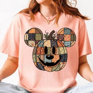 May include: A light pink t-shirt featuring a patchwork pumpkin design with Mickey Mouse ears. The pumpkin is made of various fabric patterns in orange, blue, green, and floral prints. The design has a smiling face and a stem.