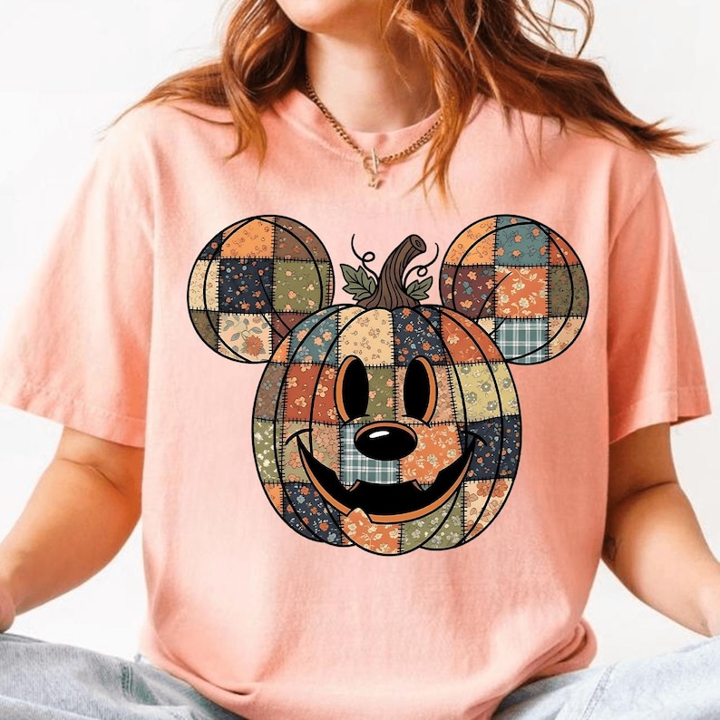 May include: A pale pink t-shirt with a Mickey Mouse pumpkin design. The pumpkin is made of patchwork fabric in orange, blue, green, and floral patterns. It has a smiling face and a stem, with Mickey Mouse ears.