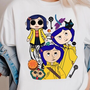May include: White t-shirt featuring a cartoon illustration of a character with blue hair and a yellow coat. The design includes a black cat, keys, and the text "Welcome Home" on a cake.