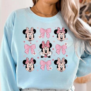 May include: Light blue sweatshirt featuring a repeating pattern of Minnie Mouse faces with pink polka dot bows and pink bows. The design includes small pink stars and white flowers, creating a playful and whimsical aesthetic.