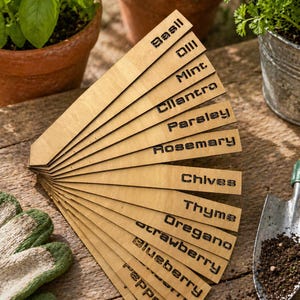 May include: A set of wooden plant markers with black engraved text. The markers are arranged in a fan shape, displaying names like Basil, Dill, and Thyme. Gardening gloves, a trowel, and potted herbs are in the background.