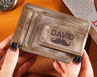 May include: A brown leather wallet with a money clip. The clip is embossed with the name "DAVID" and a mustache graphic. The wallet has several card slots and is held by a person's hands.