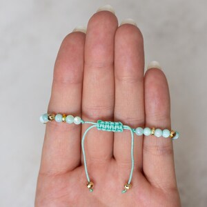 Puede incluir: Pulsera ajustable con cuentas azul claro y detalles dorados. La pulsera est&aacute; ensartada en un cord&oacute;n verde azulado claro con un cierre de nudo macram&eacute;. La pulsera se muestra en una mano.