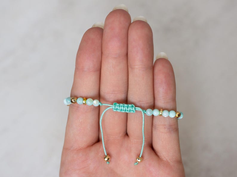 Puede incluir: Pulsera ajustable con cuentas azul claro y detalles dorados. La pulsera est&aacute; ensartada en un cord&oacute;n verde azulado claro con un cierre de nudo macram&eacute;. La pulsera se muestra en una mano.