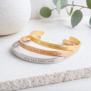 K&ouml;nnte beinhalten: Drei Metallarmreifen in Gold-, Ros&eacute;gold- und Silbert&ouml;nen. Jeder Armreif hat eine glatte, polierte Oberfl&auml;che und ist mit dem schwarzen Text THIS TOO SHALL PASS graviert. Die Armreifen sind offen gestaltet und haben ein schmales Banddesign.