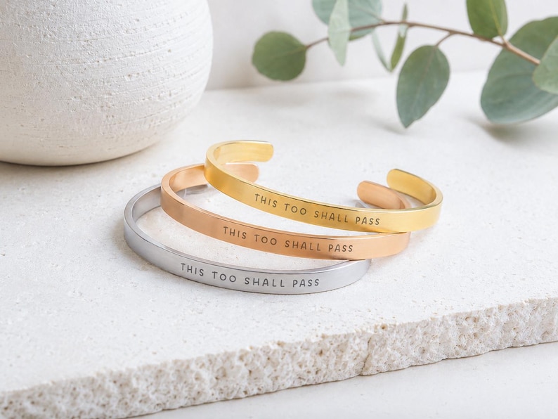 K&ouml;nnte beinhalten: Drei Metallarmreifen in Gold-, Ros&eacute;gold- und Silbert&ouml;nen. Jeder Armreif hat eine glatte, polierte Oberfl&auml;che und ist mit dem schwarzen Text THIS TOO SHALL PASS graviert. Die Armreifen sind offen gestaltet und haben ein schmales Banddesign.