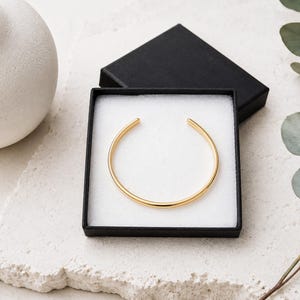 K&ouml;nnte beinhalten: Ein goldfarbenes, offenes Armband mit glatter, polierter Oberfl&auml;che. Das Armband wird in einer quadratischen schwarzen Geschenkbox mit wei&szlig;em Schaumstoffeinsatz pr&auml;sentiert und zeigt ein schlichtes, kreisf&ouml;rmiges Design mit zwei abgerundeten Enden.