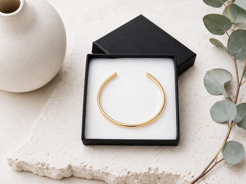 K&ouml;nnte beinhalten: Ein goldfarbenes, offenes Armband mit glatter, polierter Oberfl&auml;che. Das Armband wird in einer quadratischen schwarzen Geschenkbox mit wei&szlig;em Schaumstoffeinsatz pr&auml;sentiert und zeigt ein schlichtes, kreisf&ouml;rmiges Design mit zwei abgerundeten Enden.