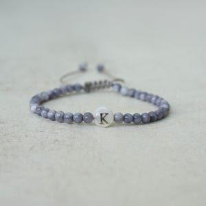 May include: A beaded bracelet with a gray and white color scheme. The bracelet features small, round beads and a central white charm with the letter "K" in the center. The bracelet is adjustable.