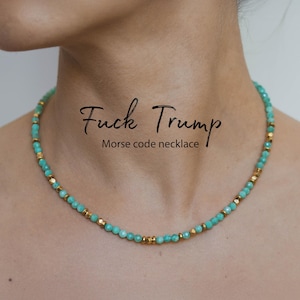 May include: A turquoise beaded necklace with gold-colored accents. The necklace is around a person's neck. The text "Fuck Trump Morse code necklace" is written in black cursive font above the necklace.