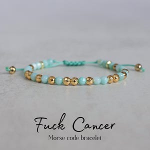 May include: A beaded bracelet with light blue and gold beads. The bracelet is strung on a light green cord and features the text "Fuck Cancer" and "Morse code bracelet".