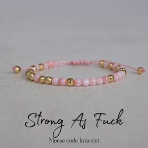 May include: Pink beaded bracelet with gold-colored accents. The bracelet features small pink beads interspersed with larger gold-colored beads. The text "Strong As Fuck" and "Morse code bracelet" are below the bracelet.