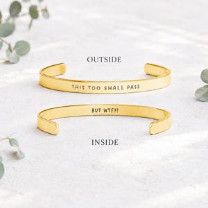 May include: Two gold-toned metal cuff bracelets. The top bracelet features the engraved text 'THIS TOO SHALL PASS' on the outside. The bottom bracelet features the engraved text 'BUT WTF?!' on the inside. Both bracelets have a smooth finish.