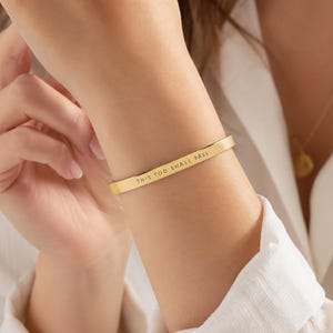 K&ouml;nnte beinhalten: Ein goldfarbenes Armband aus Metall mit glatter, polierter Oberfl&auml;che und einem flachen, schmalen Design. Die Au&szlig;enseite des Armbands ist mit dem schwarzen Text "THIS TOO SHALL PASS" in einer klaren, serifenlosen Gro&szlig;buchstabenschrift graviert.