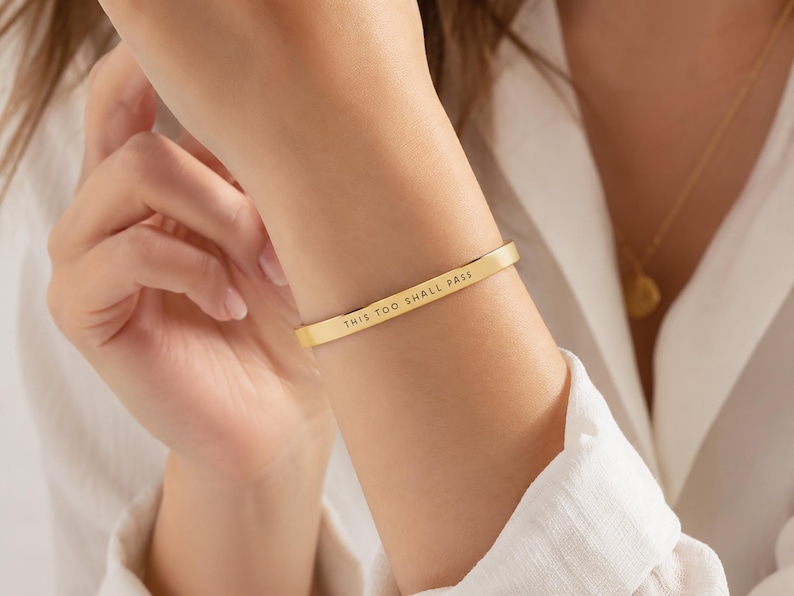 K&ouml;nnte beinhalten: Ein goldfarbenes Armband aus Metall mit glatter, polierter Oberfl&auml;che und einem flachen, schmalen Design. Die Au&szlig;enseite des Armbands ist mit dem schwarzen Text "THIS TOO SHALL PASS" in einer klaren, serifenlosen Gro&szlig;buchstabenschrift graviert.