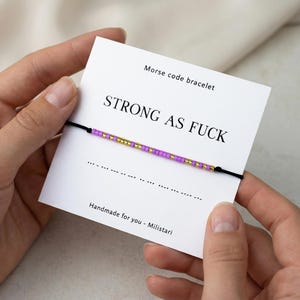 May include: A Morse code bracelet with purple and gold beads spelling "STRONG AS FUCK" on a white card. The card also says "Morse code bracelet" and "Handmade for you - Militari". The bracelet is held by two hands.