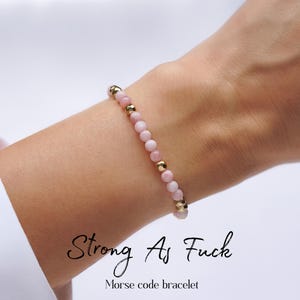 May include: A pink beaded bracelet with gold accents is worn on a wrist. The bracelet features small, round pink beads interspersed with gold-colored beads. The text "Strong As Fuck" and "Morse code bracelet" are written below the bracelet.