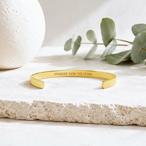 May include: A gold-toned metal cuff bracelet with an open design, featuring the engraved text STRONGER THAN THE STORM on the inner surface. The bracelet has a smooth, polished finish and rounded ends, presented in a simple, clean aesthetic.