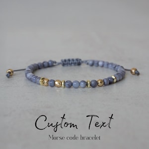 May include: A gray beaded bracelet with gold-colored accents. The bracelet features small, round gray beads interspersed with gold-colored, faceted beads. The text "Custom Text" and "Morse code bracelet" are below the bracelet.