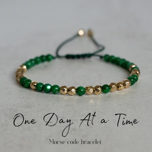 May include: Green and gold beaded bracelet with the text "One Day At a Time" and "Morse code bracelet". The bracelet features green and gold beads strung on a dark green cord, with an adjustable sliding knot closure.