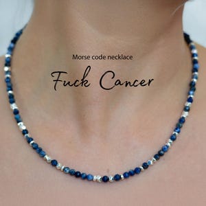 May include: A beaded necklace with dark blue and silver beads. The necklace is around a person's neck. The text "Morse code necklace" and "Fuck Cancer" are written above the necklace.