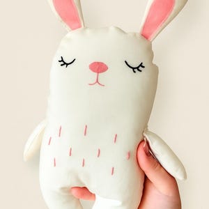 Peut inclure: Une peluche blanche en forme de lapin avec des détails roses. Le lapin a de longues oreilles avec l'intérieur rose, les yeux fermés et un nez rose. Des détails roses brodés décorent le corps. La peluche est douce.