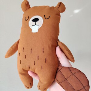 Puede incluir: Una muñeca de castor de peluche marrón hecha a mano con un diente blanco, ojos negros y una cola marrón cosida. La muñeca tiene brazos y orejas pequeños, y líneas verticales cosidas en su cuerpo. El castor se sostiene en una mano.
