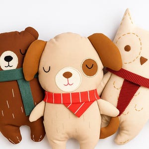 May include: Three plush animal pillows: a brown bear with a green scarf, a tan dog with a red scarf, and an off-white owl with a red scarf. Each pillow has stitched details and a whimsical design, perfect for a nursery or children's room.