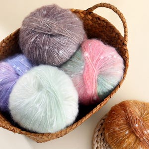 May include: A wicker basket containing several balls of colourful yarn. The yarn is in shades of lilac, pink, mint green, and brown, with white spots. The basket has a handle and is made of a light brown material. The yarn appears soft and fluffy.
