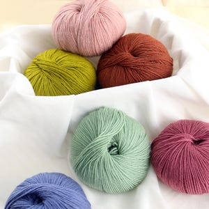 Puede incluir: Una colección de ovillos de lana coloridos en tonos rosa, verde, azul y marrón, dispuestos sobre una tela blanca. La lana parece suave y esponjosa, ideal para proyectos de tejido o ganchillo. Los colores varían de claros a oscuros, ofreciendo diversas opciones para crear.