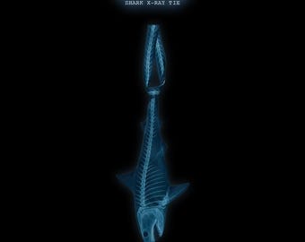 Könnte beinhalten: Eine Krawatte mit einer bläulichen Röntgenaufnahme eines Haiskeletts vor einem schwarzen Hintergrund. Der Text TIE A KNOT und SHARK X-RAY TIE ist in Weiß über der Haifischgrafik gedruckt, die oben zu einem Knoten gebunden ist.