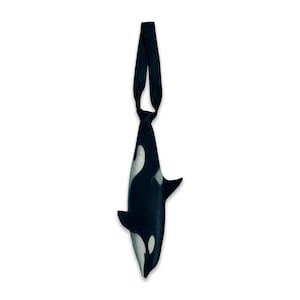 Puede incluir: Una corbata con forma de orca, que presenta el característico patrón de color negro y blanco del mamífero marino. La corbata incluye un nudo en la parte superior y se estrecha hacia el cuerpo de la ballena, con aletas laterales extendidas.