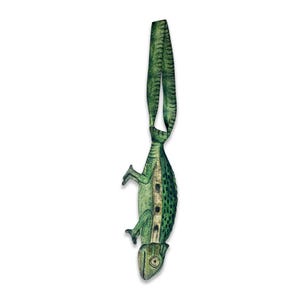 Puede incluir: Un adorno colgante con forma de camaleón verde que presenta una superficie texturizada con manchas de color verde oscuro, una franja central clara y un lazo en la parte superior para colgar. La figura del lagarto se muestra en vertical.