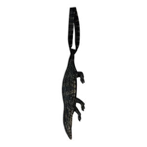 May include: A black necktie featuring a textured, reptilian-inspired pattern with subtle gold-toned highlights. The tie is shaped like an elongated alligator or crocodile, with a long body, four distinct legs, and a pointed tail at the bottom end.