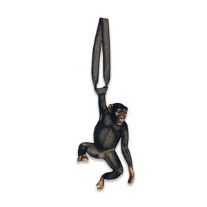 May include: A wooden chimpanzee ornament, hanging from a dark gray loop. The chimpanzee is depicted in a realistic style, with detailed fur and facial features. The ornament is designed to hang and is set against a white background.