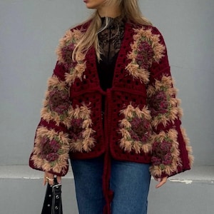 May include: A burgundy crochet cardigan with square patterns and fluffy accents in shades of tan, brown, and green. The open-front design features a tie closure and long sleeves. The cardigan is paired with blue jeans.