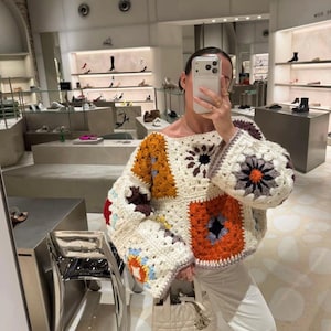 May include: A cream-coloured, patchwork crochet jumper with square patterns in orange, brown, and blue. The jumper has wide sleeves and a round neckline. The image is taken in a shop with shoes on display in the background.
