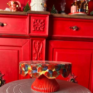 Może przedstawiać: Czerwony ceramiczny stojak na ciasto z falistą krawędzią i podstawą. Górna powierzchnia jest ozdobiona kolorowymi pociągnięciami pędzla w odcieniach czerwieni, niebieskiego i żółtego. Dekoracyjny element do prezentacji ciast.