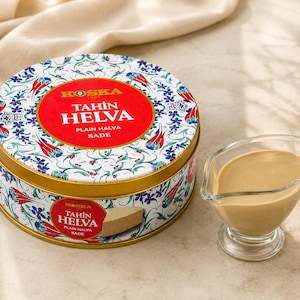 Puede incluir: Una lata redonda de Tahin Helva con un diseño floral rojo y blanco, y un pequeño plato de vidrio lleno de una salsa marrón claro. La lata tiene un borde dorado y el texto "KOSKA TAHIN HELVA PLAIN HALVA SADE".