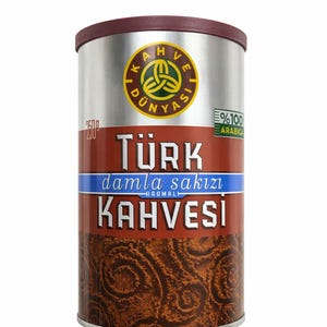 May include: A cylindrical coffee tin with a maroon lid and a silver body. The tin features Turkish text and a logo with a green and yellow design. The bottom half of the tin has a brown, textured pattern.