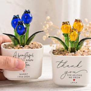Puede incluir: Dos pequeñas macetas de cerámica blanca con esculturas de flores de cristal. Una maceta tiene flores azules, la otra flores amarillas. Las macetas tienen el texto "A beautiful life never ten" y "thank you for helping me grow".