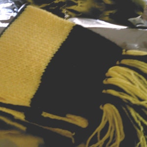 以下が含まれることがあります： フリンジが付いた黒と黄色のニットスカーフ。スカーフは半分に折り畳まれており、片側に黄色のストライプがあります。