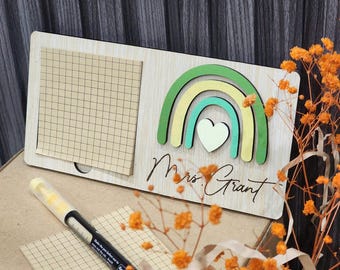 May include: Wooden desk organizer with a rainbow design in green, yellow, and teal, and a heart. The organizer has a notepad and the name "Mrs. Grant" engraved on it. A pen and grid paper are also visible.