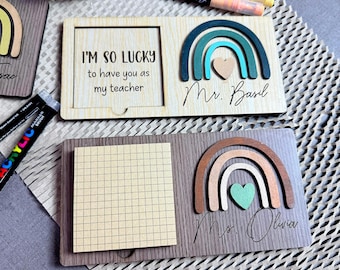 May include: Wooden teacher appreciation gifts with rainbow and heart accents. One has a frame with the text "I'M SO LUCKY to have you as my teacher" and "Mr. Basil". The other has a notepad and "Ms. Olivia".