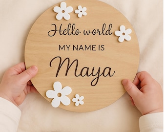 Peut inclure: Un panneau rond en bois avec le texte "Hello world, MY NAME IS Maya" en écriture noire. Le panneau est orné de fleurs blanches. Le panneau est tenu par une personne.