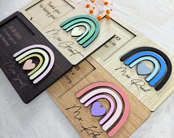May include: Wooden rainbow-themed teacher appreciation gifts. Each features a colorful rainbow and heart design, with personalized text like "Mrs. Grant" and "Thank you for being you!" on a wooden plaque. The color palette includes pastels and natural wood tones.