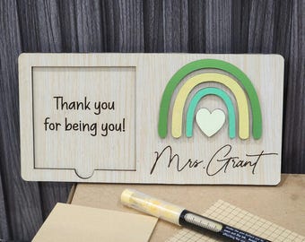 May include: Wooden plaque with a cutout for a photo and a colorful rainbow design. The text "Thank you for being you!" is on the left, and "Mrs. Grant" is written below the rainbow. A yellow pen and notepad are in the foreground.