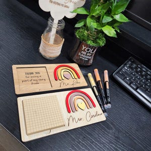 Op de afbeelding: Houten cadeaus voor lerarenwaardering met regenboogontwerpen. Eén heeft ruimte voor een foto en de tekst "THANK YOU for being a part of my story, Brooke." De andere heeft een geruit notitieblok. Pennen en een plant zijn ook zichtbaar.